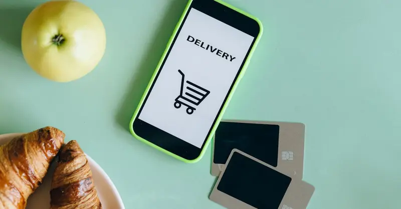 A smartphone with a delivery app next to an apple, croissants, and credit cards on a green surface.
