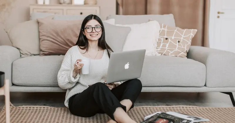 Full body happy young female in casual clothes sitting on floor near sofa with cup of tea and laptop while surfing Internet