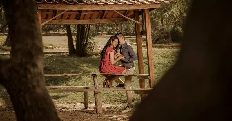 A loving couple sitting on a picnic table, sharing a tender moment under a roofed structure in a serene outdoor setting.