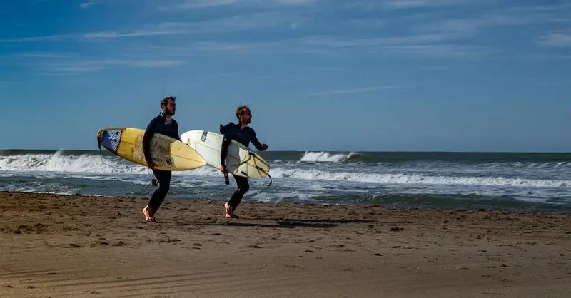 Two surfers with boards walk along Cariló beach in Argentina under a clear blue sky.