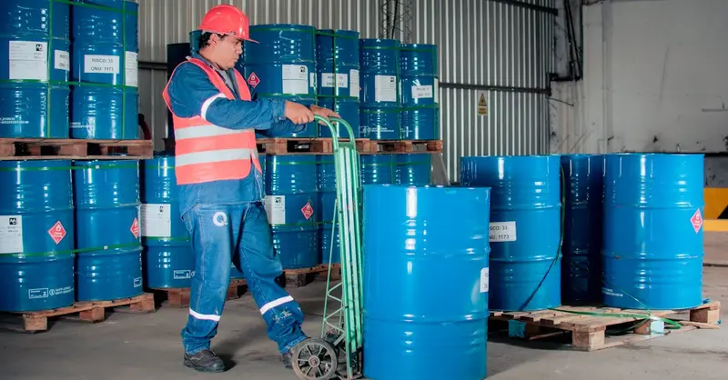 Industrial worker with a hand truck moving blue barrels in a warehouse setting.