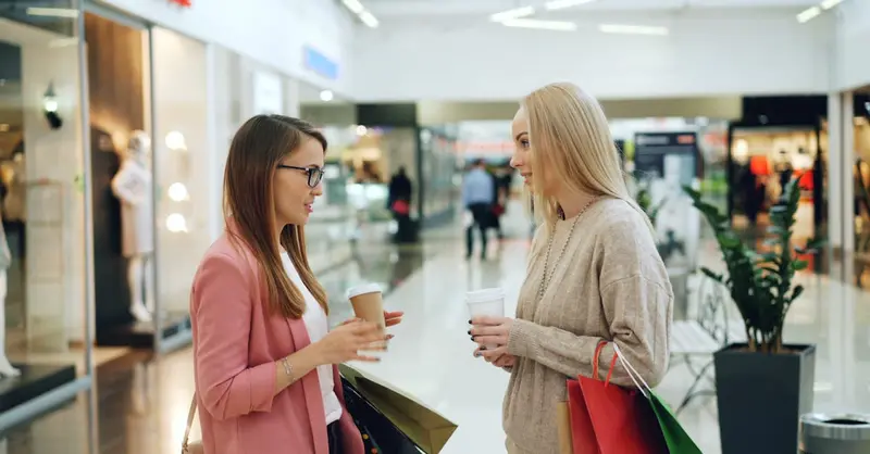 Two women conversing in a shopping mall holding coffee and shopping bags.