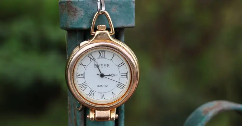 A vintage pocket watch with Roman numerals hanging on a rustic green metal fence outdoors.