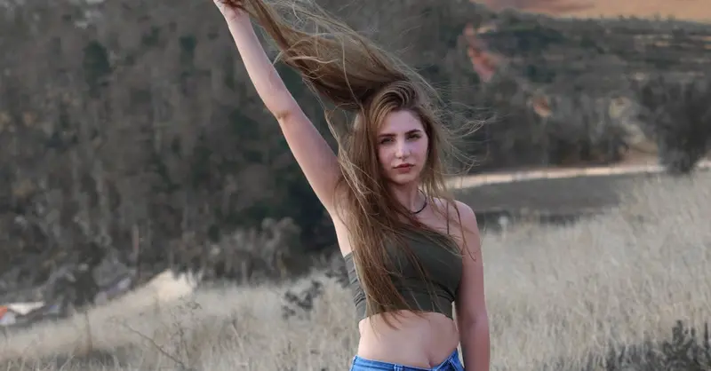 A young woman in a tube top and jeans poses in a rural Brazilian landscape, holding her hair aloft for a dynamic effect.