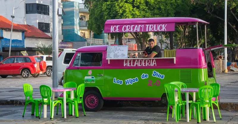 Bright ice cream truck serving customers on a sunlit urban street with colorful chairs.