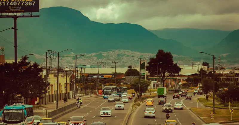 Traffic flows on a busy urban road with a mountainous backdrop under cloudy skies.