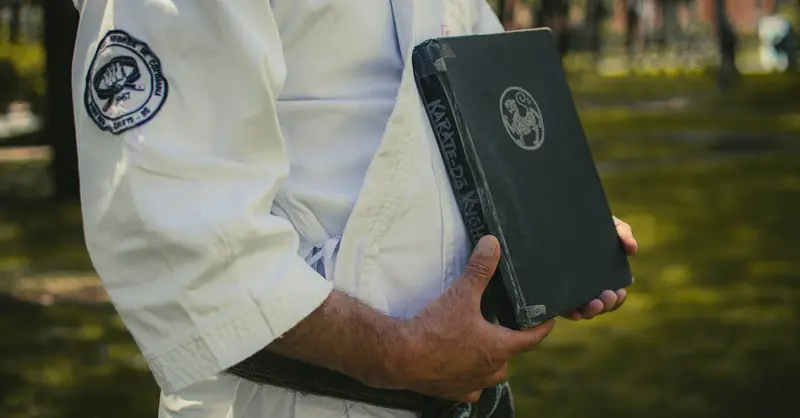 Close-up shot of a martial artist in a karategi holding a black karate reference book outdoors.