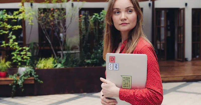 A young woman in a red dress holding a laptop outdoors, looking thoughtful.