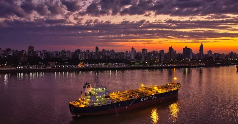 A cargo ship navigates a calm river at sunset, with a vibrant city skyline backdrop.