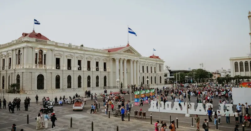Crowded plaza scene in San Salvador with flags and historic buildings under a clear sky.