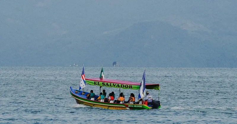 A colorful boat with flags sails on a serene lake in El Salvador, surrounded by scenic mountains.