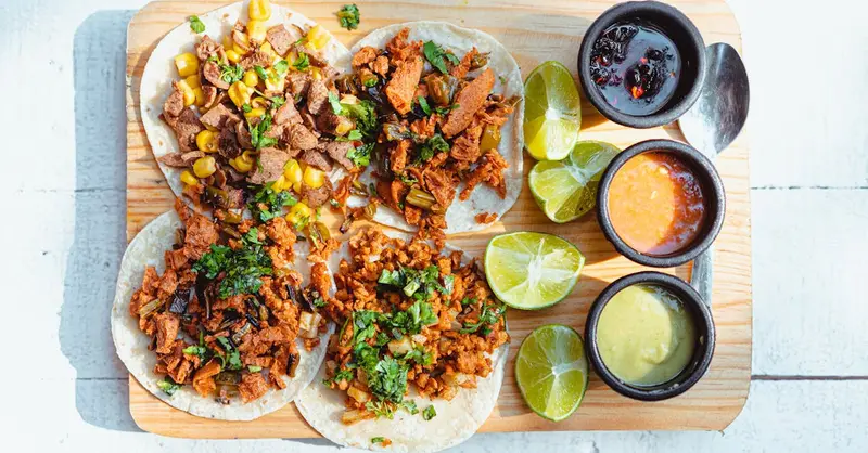 Colorful vegan tacos with lime slices and dipping sauces on a wooden tray, shot in bright light.