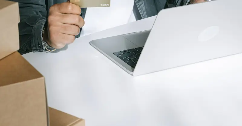 Person using a credit card for online shopping on a laptop surrounded by shipping boxes.