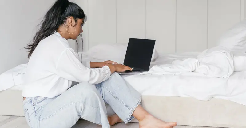 A woman sits on the floor working on a laptop in a serene bedroom environment, embodying remote work and relaxation.