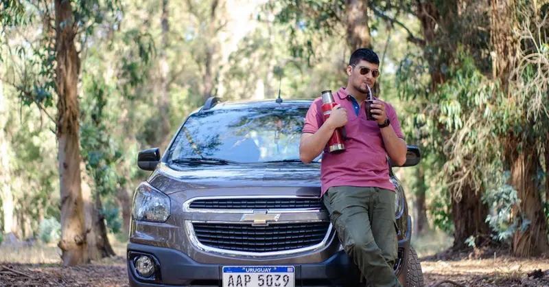 Man leaning on a Chevrolet in a forest in Uruguay, enjoying a drink with a thermos.