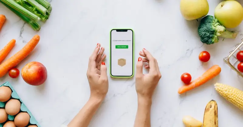 Hands holding smartphone displaying online grocery order amidst fresh vegetables and fruits.