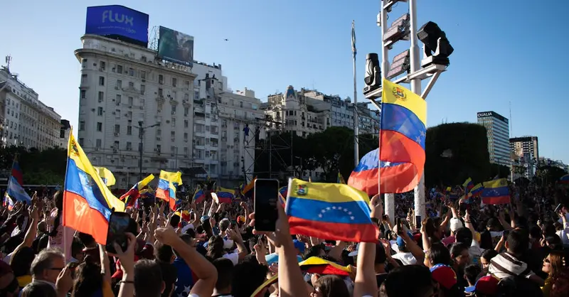 A large crowd waves Venezuelan flags during a protest in a bustling city square.