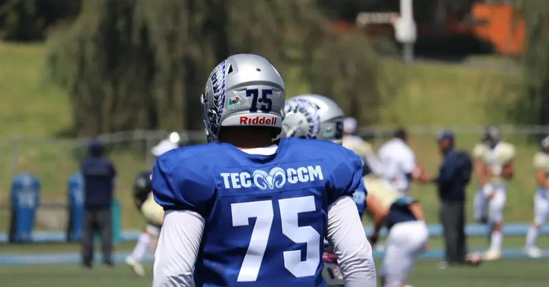 A college football player in uniform on the field, preparing for the game with teammates in the background.